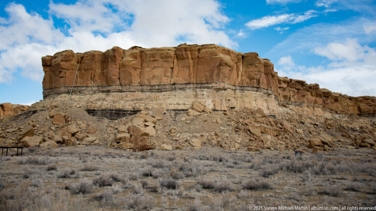 A layered sandstone cliff with a reddish cap rises above pale eroded rock and a talus-covered slope.