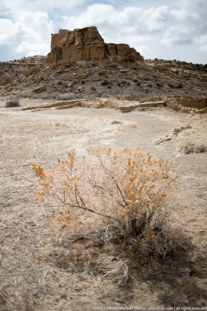 Low stone remains sit on a rocky ridge above a pale open slope dotted with dry brush.