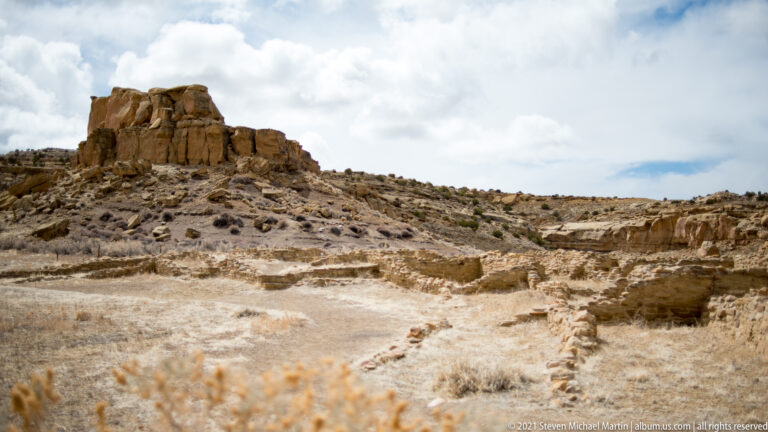 Low stone walls and room outlines extend below a blocky sandstone outcrop and canyon cliffs.