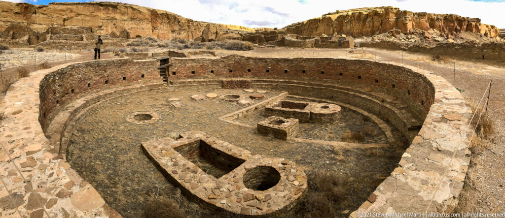 A visitor stands beside a large circular excavated structure with interior masonry features and cliffs beyond.