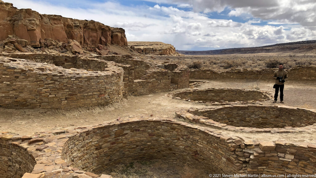 A visitor stands among low masonry walls and excavated spaces, with the canyon opening in the distance.