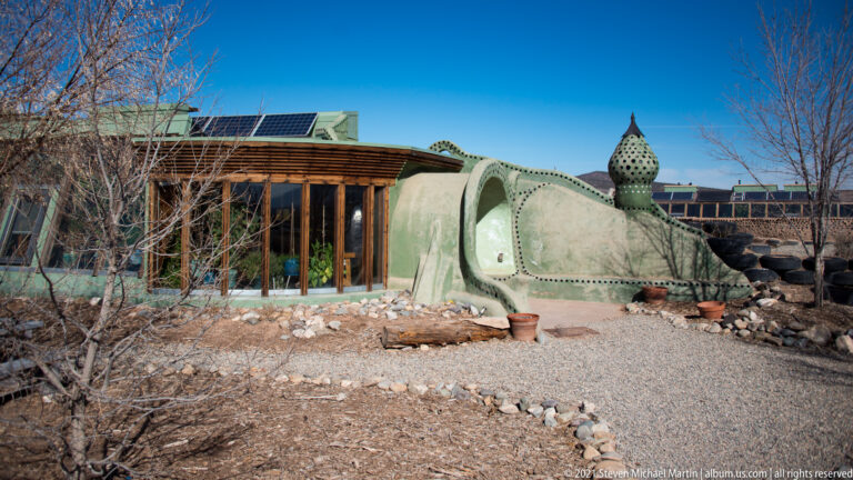 A curved earth-toned residence has a glazed front, rooftop solar panels, a sculptural vent, and gravel landscaping.