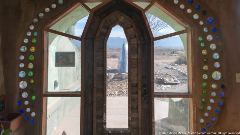 A tall glazed doorway is framed by earth-colored walls embedded with rows of colored glass bottles.