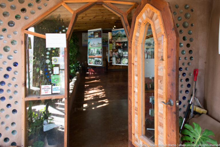 Open wooden doors lead into a plant-filled gallery through walls set with colored glass bottle ends.