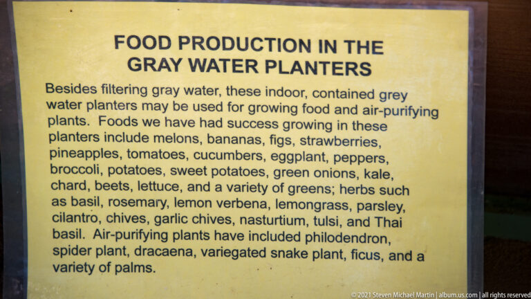 A yellow sign lists vegetables, herbs, and foliage plants grown in indoor graywater planters.