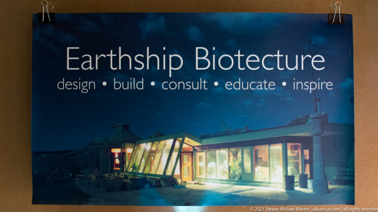 An Earthship Biotecture poster shows a warmly lit residence at night beneath the organization’s name and services.