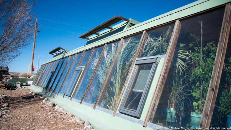 Angled wood-framed windows extend along a low building, revealing plants inside and a small openable pane.