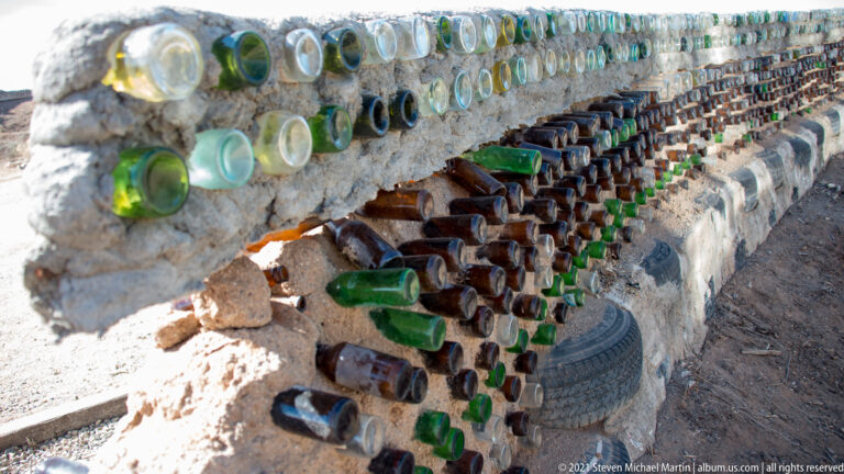 Colored glass bottle ends are embedded in a rough wall above dark tires and stones.