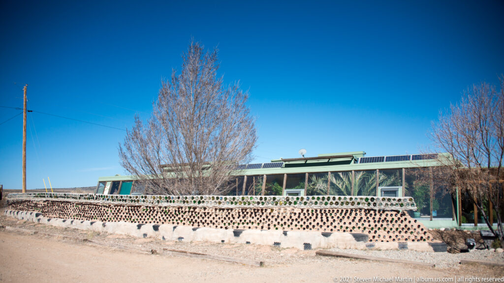 A low glazed building stands behind a curving wall patterned with bottle ends, with leafless trees nearby.