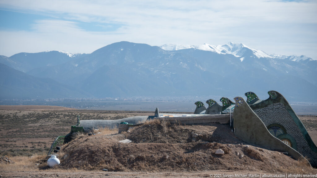 A low earth-covered mound and bottle-studded wall sit on a plain below distant snow-dusted mountains.