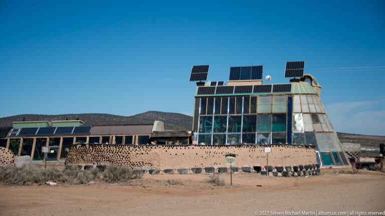 A low earthen building with a tall glazed section and solar panels stands behind a bottle-studded wall.
