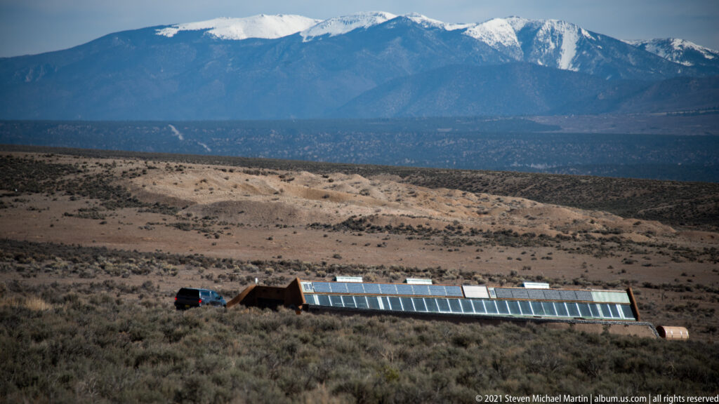 A long glazed façade and low earth-covered building sit on an open plain beneath distant mountains.