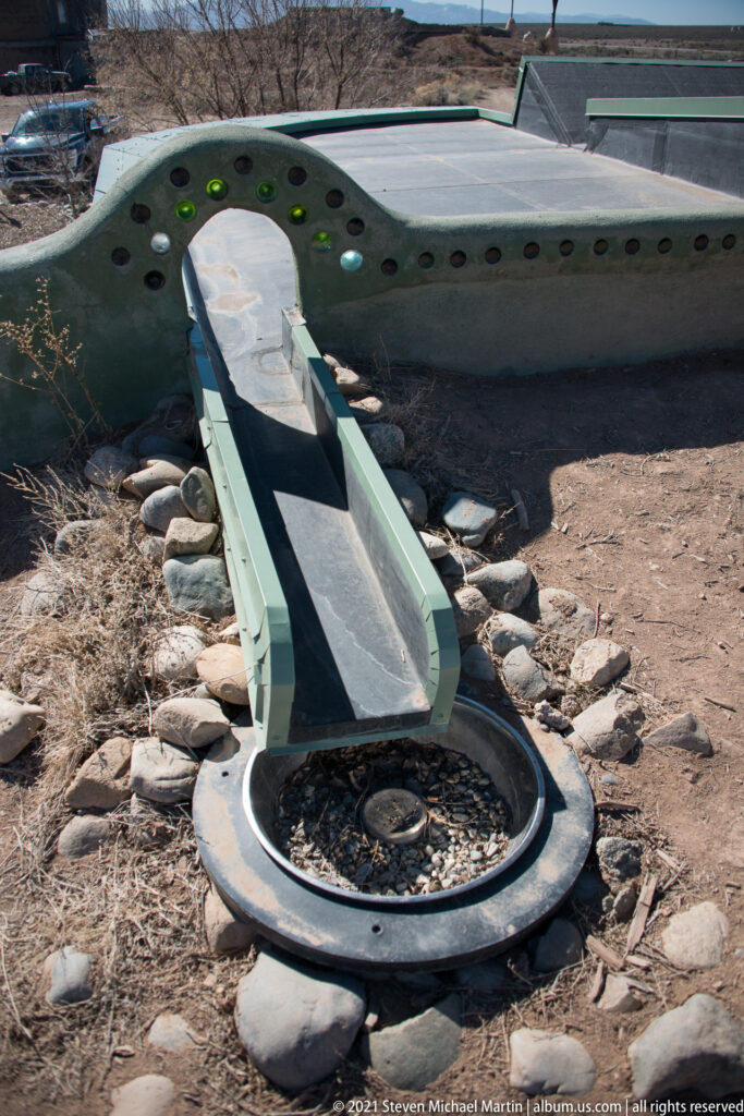 A narrow metal channel extends from a green sculpted wall toward a shallow round basin among stones.