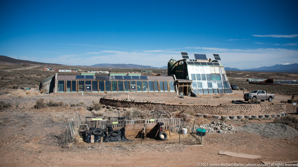 A long solar-equipped glass building stands beyond a yard containing tires and materials, with mountains behind.