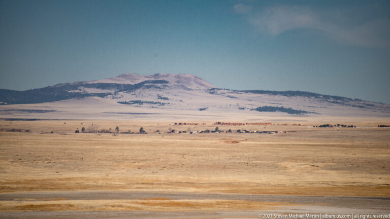 A snow-dusted mountain rises beyond a broad tan plain and a thin line of trees.