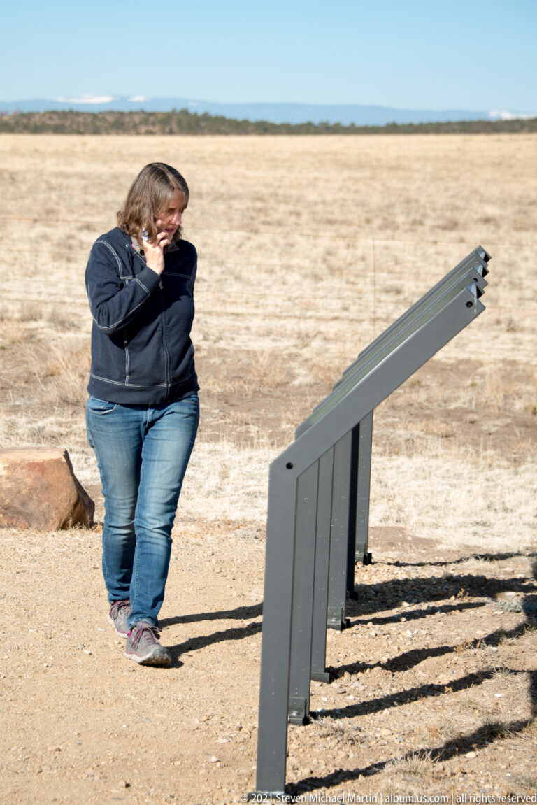 A woman walks beside a low angled metal sign on a sandy path through dry prairie.