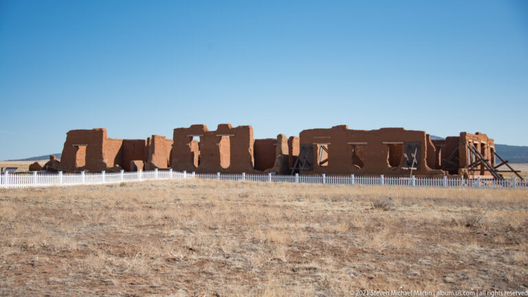 Roofless adobe rooms with broken parapets stand behind a low white fence on open grassland.