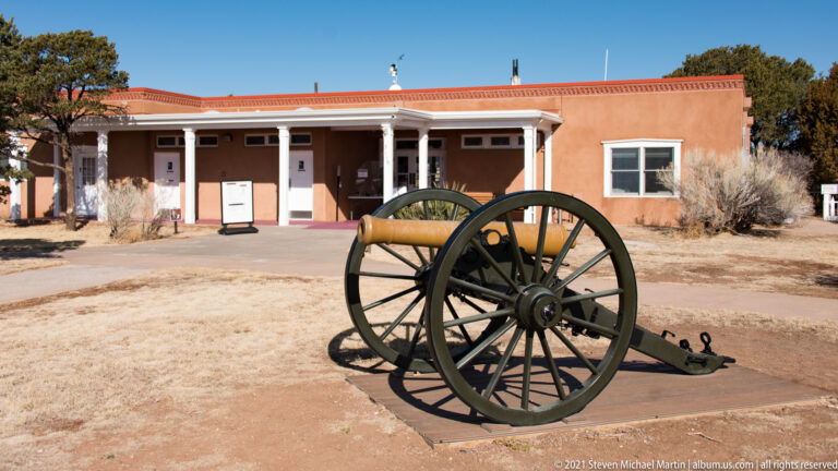 A black wheeled cannon stands on a paved pad before a low adobe-colored building.