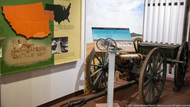Field cannon on a wooden carriage stands before a large interpretive map in a museum exhibit.