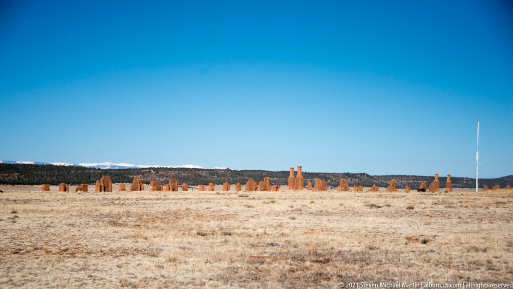 Low adobe ruins stretch across a grassy plain, with a distant tree line beneath an open sky.
