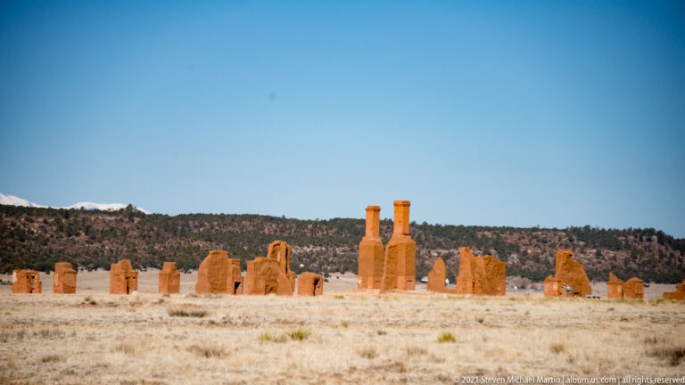 Two tall chimneys rise above low adobe walls and grassy ground in a broad view of Fort Union ruins.