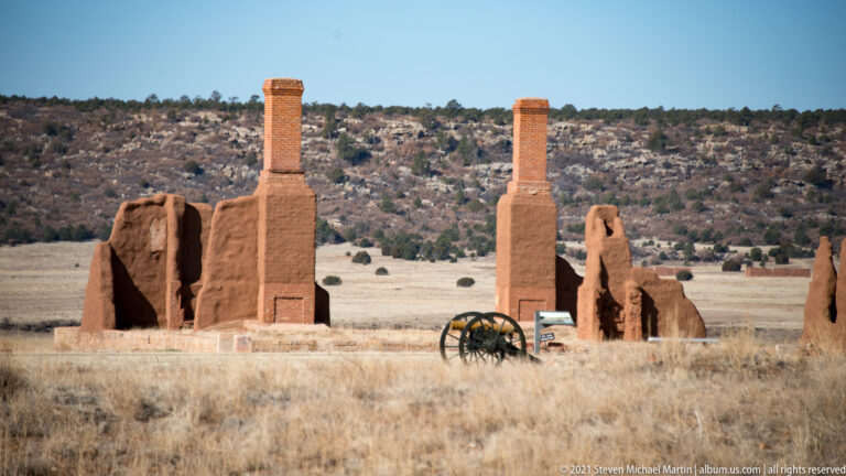 A cannon stands before adobe ruins with two tall chimneys and a wooded ridge beyond.