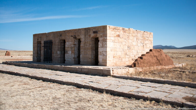 Low stone building stands on a raised platform beside a broken masonry wall and walkway.