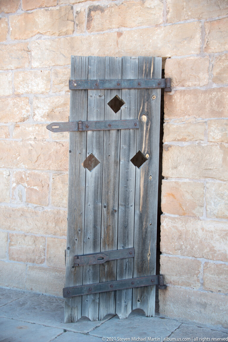 Small gray shutter with diamond openings is inset in rough masonry, shown in close view.