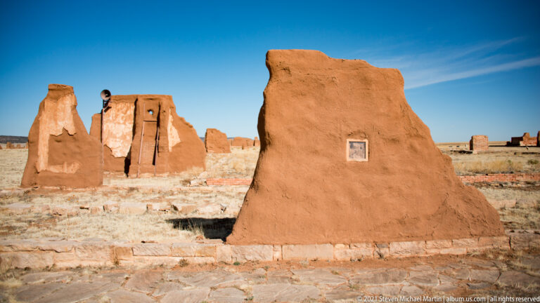 Large weathered adobe wall with a small plaque stands in front of additional ruins.