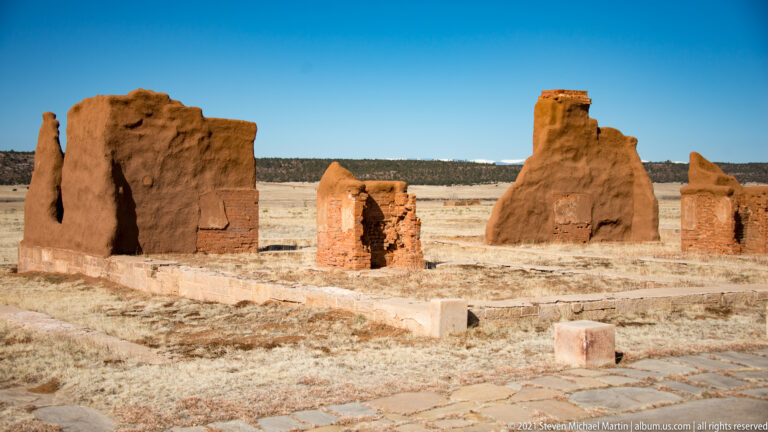 Adobe wall and chimney remnants rise from low foundations in a grassy open area.