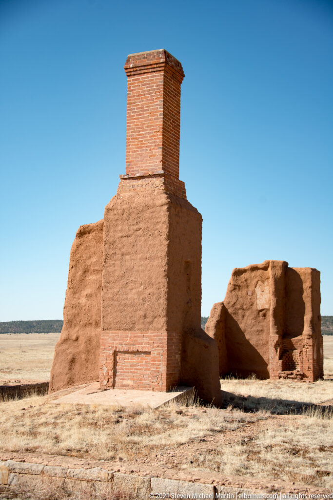 Tall brick chimney rises above a broad adobe wall, with lower ruin fragments nearby.