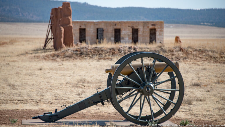 Field cannon stands on open ground before a long stone and brick building.