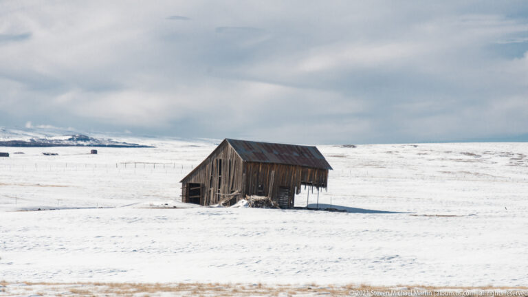 A small weathered wooden outbuilding stands alone in a snowy field beneath gray clouds.
