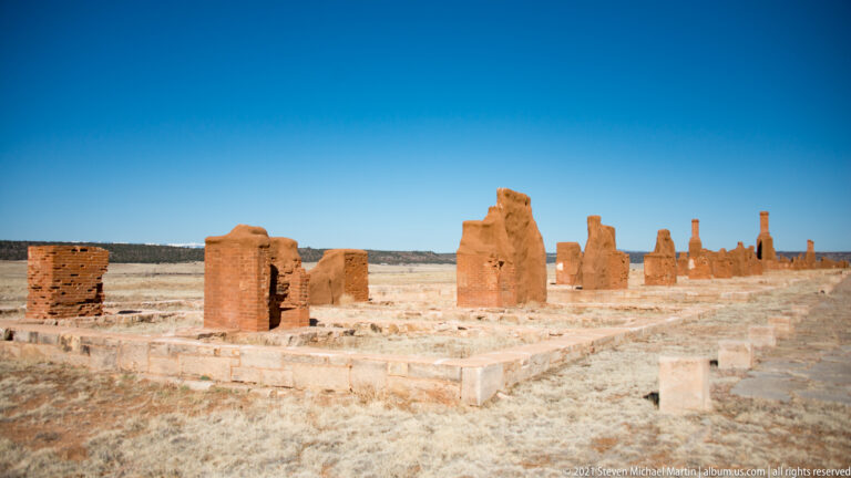 Low adobe foundations extend across an open site beside a path in the right foreground.