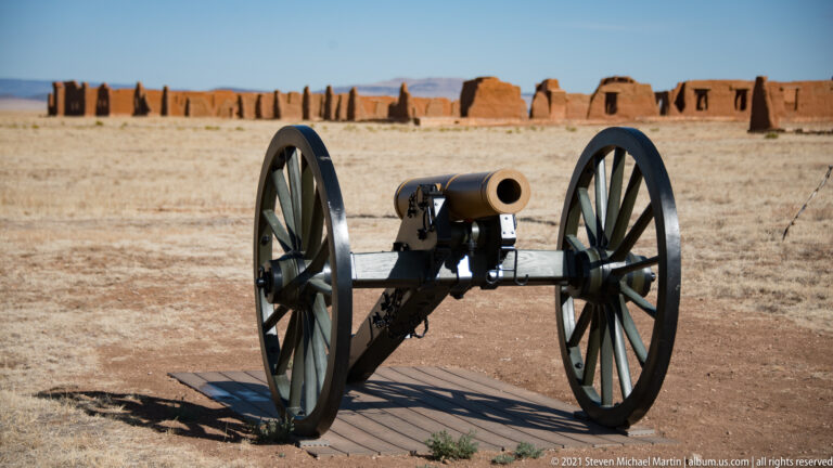 Cannon muzzle faces forward between large wheels, with adobe ruins and a chimney behind it.