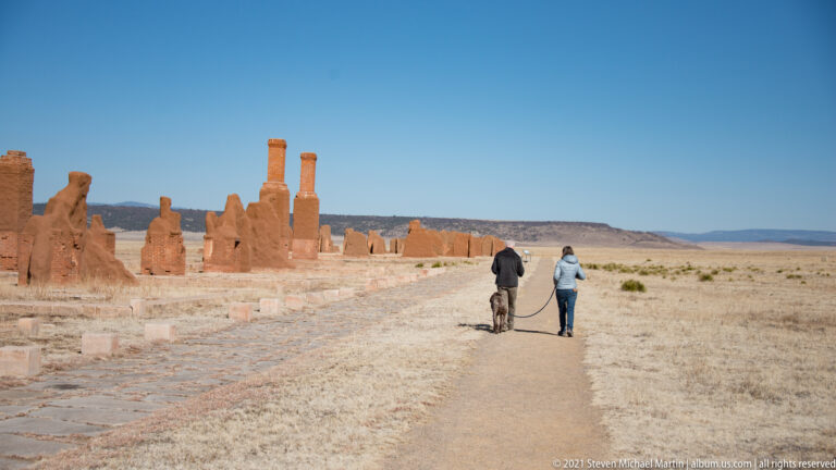 Two visitors walk along a path beside low adobe ruins in an open grassy landscape.