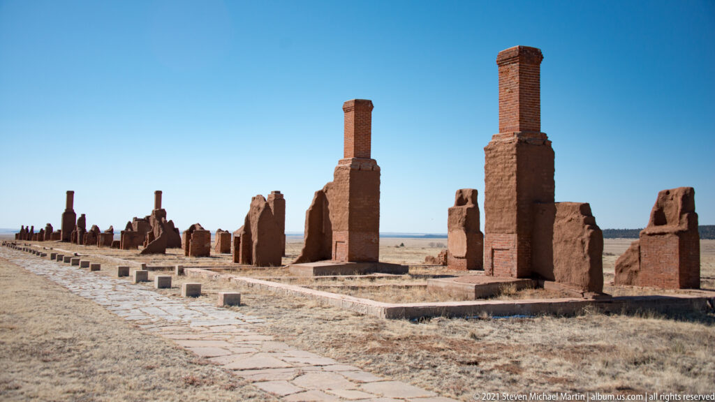 Paired chimneys rise above adobe ruins beside a stone path running diagonally through the scene.