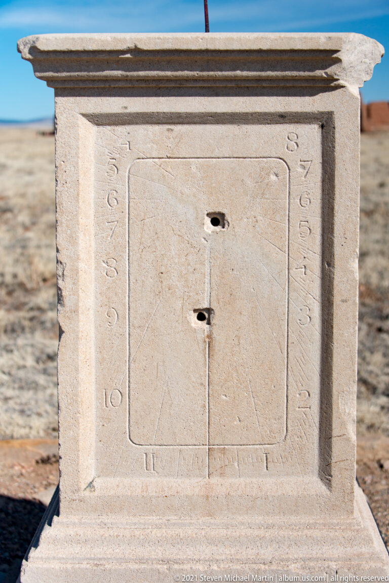 Front view of a pale stone sundial pedestal with Roman numerals, dark central fittings, and a narrow desert backdrop.