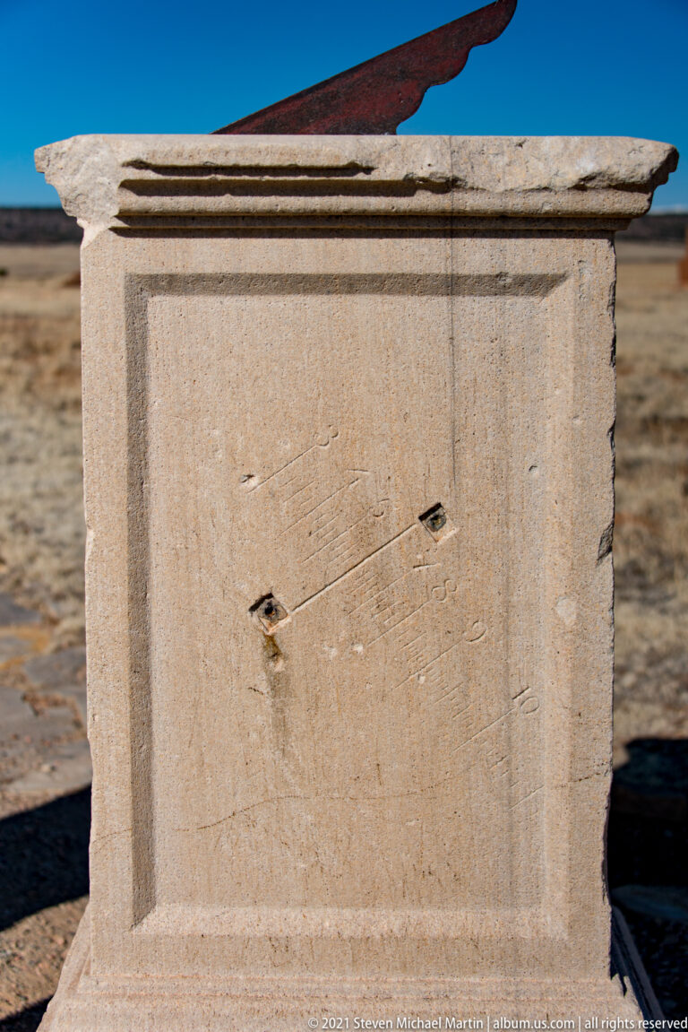 Plain reverse face of a pale stone pedestal, with two dark fittings and a projecting cap against dry grass.