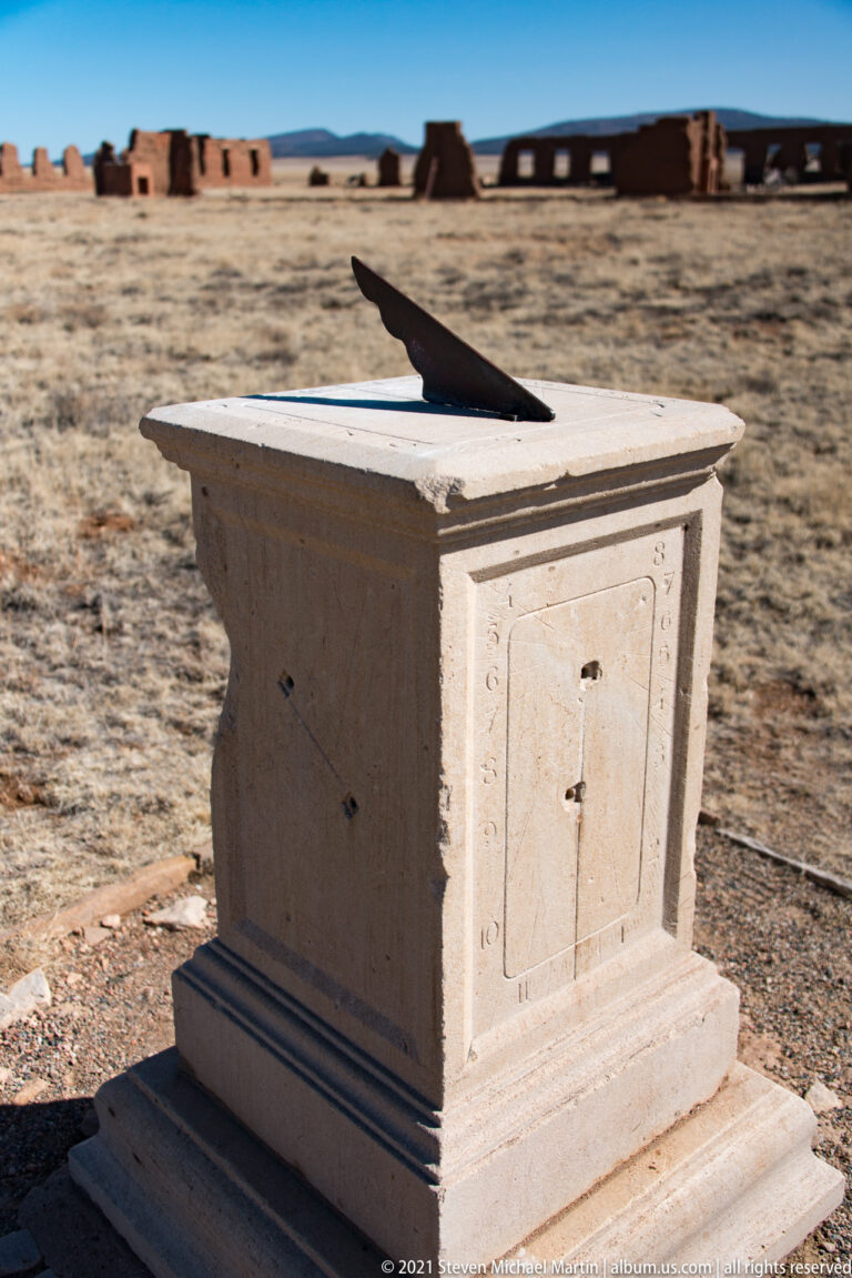 Three-quarter view of a stepped stone sundial pedestal with a dark triangular piece on top and adobe ruins behind.