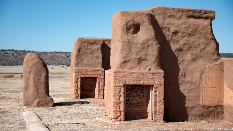 Two eroded adobe structures with brick-lined doorways stand in dry grass beneath a clear sky.