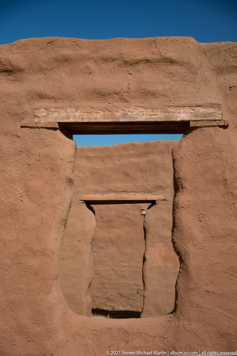 Close view of two narrow openings with timber lintels in a broad, weathered adobe wall.