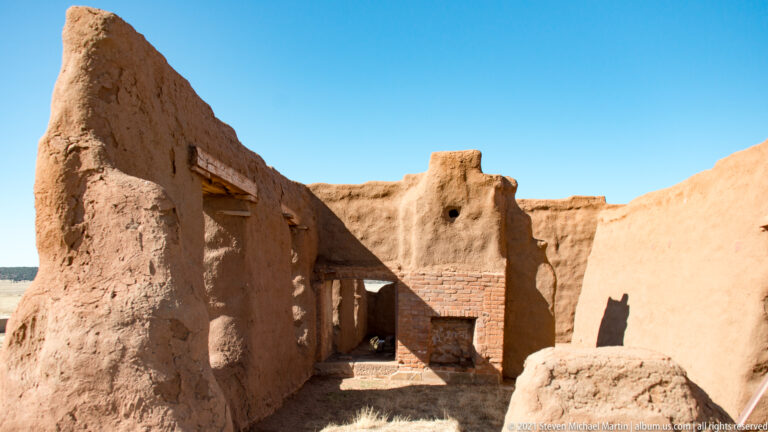 Sunlit adobe courtyard with brick-edged doorways, irregular walls, and deep shadows between structures.