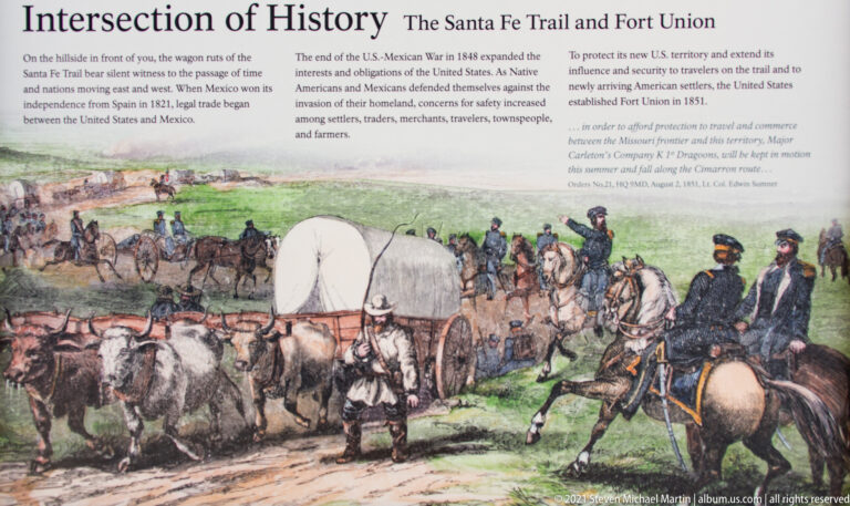 A painted wagon train with covered wagons, draft animals, and riders crosses a grassy plain.