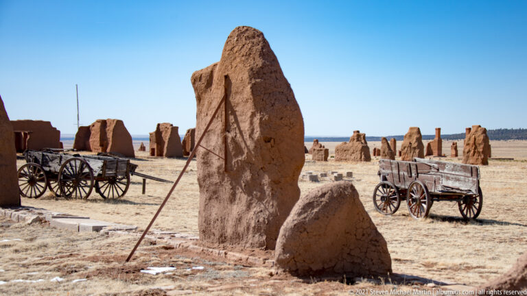 Two wooden wagons among low adobe ruins, with a tall earthen remnant rising in the foreground.