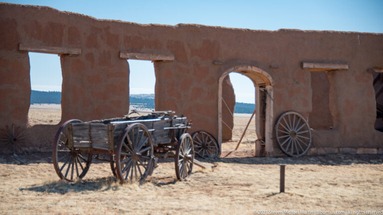 Weathered wooden wagon in the shade of an adobe gateway, with a loose wheel leaning against the wall.