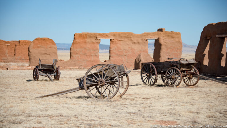 Multiple wooden wagons, including a wheel-and-shaft frame, stand in an open yard among adobe walls.