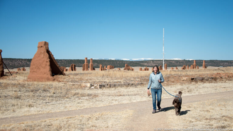 A visitor walks a leashed dog across open grassland, with scattered adobe ruins and a tall wall remnant behind.