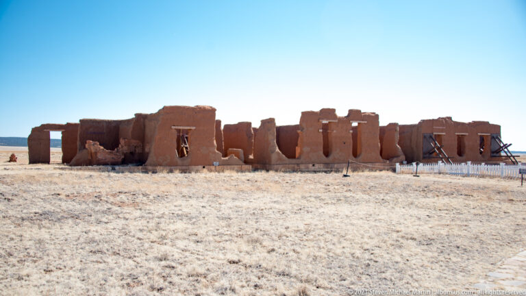 Distant view of a long roofless adobe building with repeated openings and timber braces supporting weakened walls.