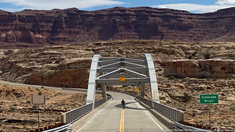 A two-lane bridge approaches the canyon beneath layered cliffs, with a Colorado River sign at the roadside.